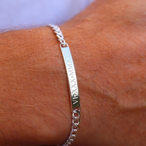 May include: A silver curb chain bracelet with a rectangular bar. The bar is engraved with Roman numerals. The bracelet is worn on a person's wrist.