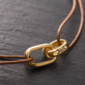 May include: A gold-colored necklace featuring two interlocking links. One link is a rounded rectangle, and the other is a ring engraved with the name "Jack." The necklace is strung on a brown cord, set against a dark background.