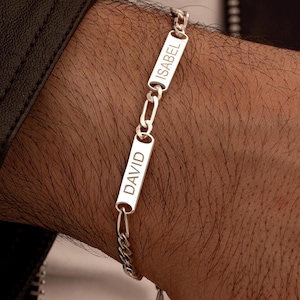 Puede incluir: Una pulsera de plata con dos placas rectangulares con los nombres "DAVID" e "ISABEL". La pulsera está unida con un diseño de cadena y se lleva en la muñeca. El fondo es una chaqueta de cuero oscura.