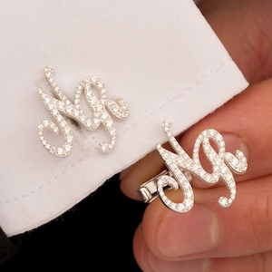 Puede incluir: Un par de gemelos de plata con un diseño engastado en diamantes. Los gemelos tienen la forma de las letras "NA".