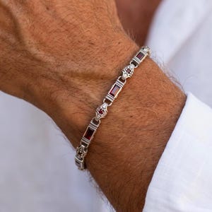 Puede incluir: Una pulsera de eslabones de plata con piedras de granate rojo oscuro rectangulares que se alternan con pequeños segmentos de plata ornamentados. La pulsera tiene un acabado metálico pulido con detalles intrincados en los conectores.