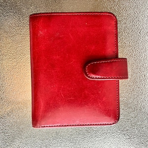 May include: A vibrant red leather planner with a matching tab closure. The planner is rectangular with rounded corners and a stitched border. The leather has a slightly distressed appearance.