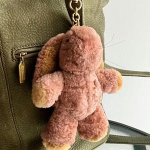 May include: A plush pink bunny keychain with tan accents on the ears and paws. The bunny is attached to a gold-tone metal clasp, hanging from a green leather bag. The bunny has a textured, fuzzy appearance and small pink details.