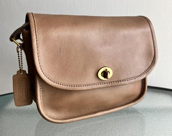 May include: A tan leather crossbody bag with a flap closure and a gold-tone turn lock. The bag has a stitched design and a small leather tag with a gold chain. The bag is a vintage style.