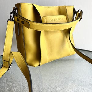 May include: A yellow leather handbag with a shoulder strap and a top handle. The bag has a rectangular shape with a flap and the brand name "LONGCHAMP" embossed on the front. The bag is sitting on a glass surface.