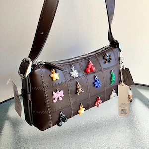 Puede incluir: Un bolso de hombro de cuero marrón con un diseño de patchwork. El bolso tiene una cremallera plateada y una correa larga y ajustable. La parte delantera está decorada con abalorios de colores en varias formas, incluyendo estrellas, flores y lazos. Una etiqueta cuelga del lateral.