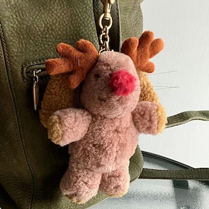 May include: A plush reindeer keychain in pink, orange, and tan. The reindeer has a red nose and small black whiskers. The keychain is attached to a gold-colored clasp and chain, hanging from a green leather bag.