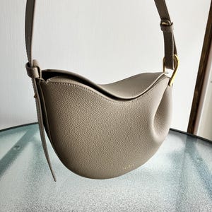 May include: A taupe-colored, crescent-shaped handbag with a long shoulder strap. The bag has a textured surface and a gold-colored metal ring connecting the strap. The design is minimalist and modern, suitable for everyday use.