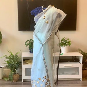 May include: A light blue sheer saree with gold and black embellishments. The saree has a black border and a white trim. The saree is draped over a mannequin.