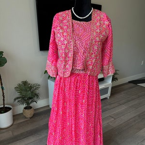 May include: A pink and gold three-piece Indian outfit with a long skirt, a short-sleeved top, and a matching jacket. The outfit is decorated with intricate patterns and sequins.