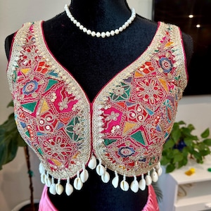 May include: A pink and gold embroidered crop top with a white shell trim and a pearl necklace. The top is on a black mannequin.