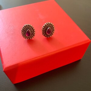 May include: A pair of stud earrings with a dark metal finish. Each earring features a teardrop-shaped, deep red gemstone set within a beaded, circular frame. The earrings are displayed on a bright red box.