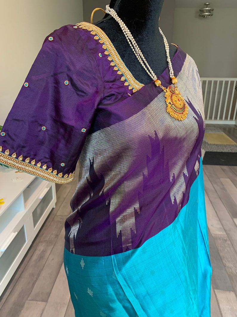May include: A purple and turquoise silk saree with gold embroidery and a pearl necklace. The saree has a silver and purple patterned design.