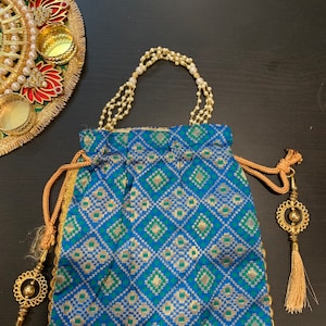 Puede incluir: Una bolsa de cordón azul y verde con estampado con un asa de cuentas doradas y detalles de borlas doradas.