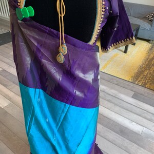 May include: A purple and blue silk saree with gold embroidery. The saree is draped over a mannequin and has a gold tassel hanging from the back.