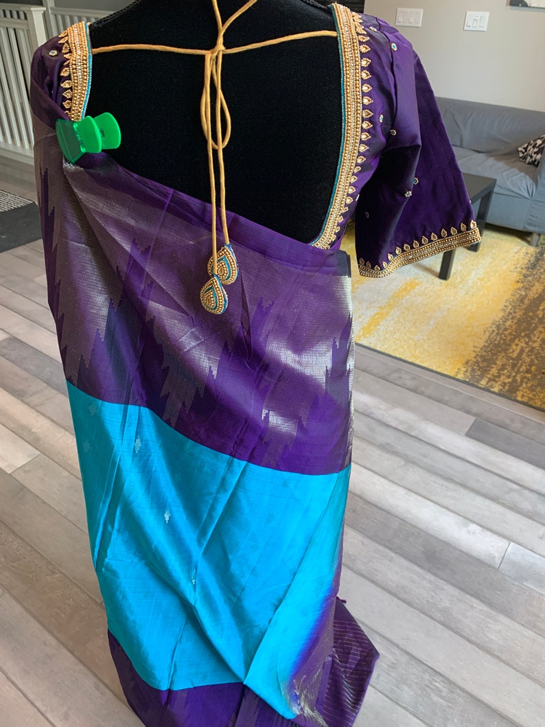May include: A purple and blue silk saree with gold embroidery. The saree is draped over a mannequin and has a gold tassel hanging from the back.