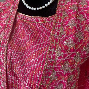 May include: A pink and gold embroidered top with a pearl necklace. The top has a floral pattern and sequins.