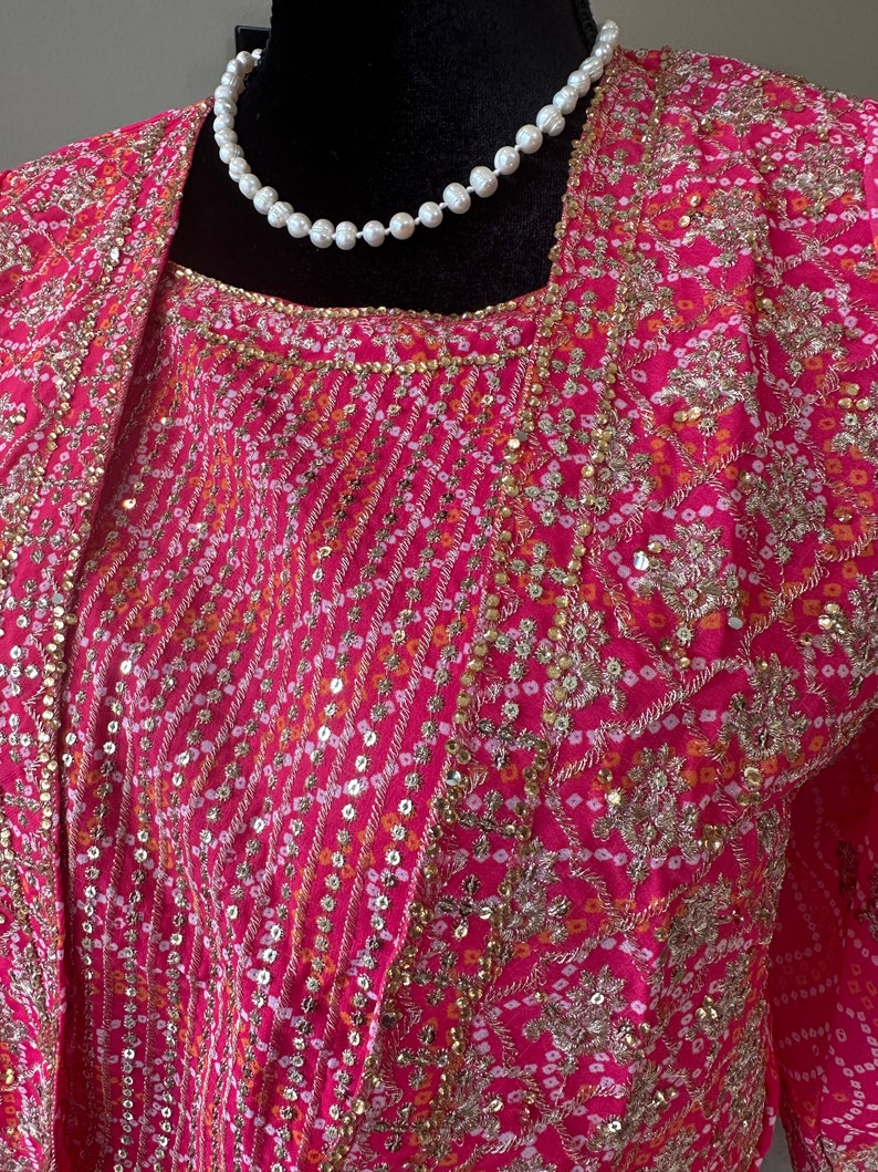 May include: A pink and gold embroidered top with a pearl necklace. The top has a floral pattern and sequins.