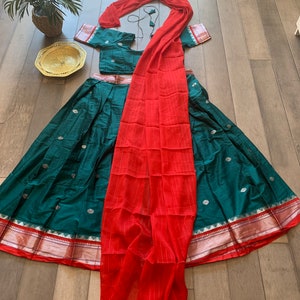 May include: A teal green skirt with gold and silver accents, a red dupatta, and a matching teal green blouse. The skirt has a gold and silver border and the dupatta is sheer with a striped pattern.