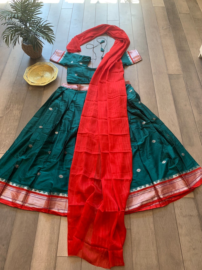 May include: A teal green skirt with gold and silver accents, a red dupatta, and a matching teal green blouse. The skirt has a gold and silver border and the dupatta is sheer with a striped pattern.