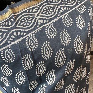 May include: A black cotton scarf with a white paisley pattern and gold trim. The scarf is folded and draped over a black surface.