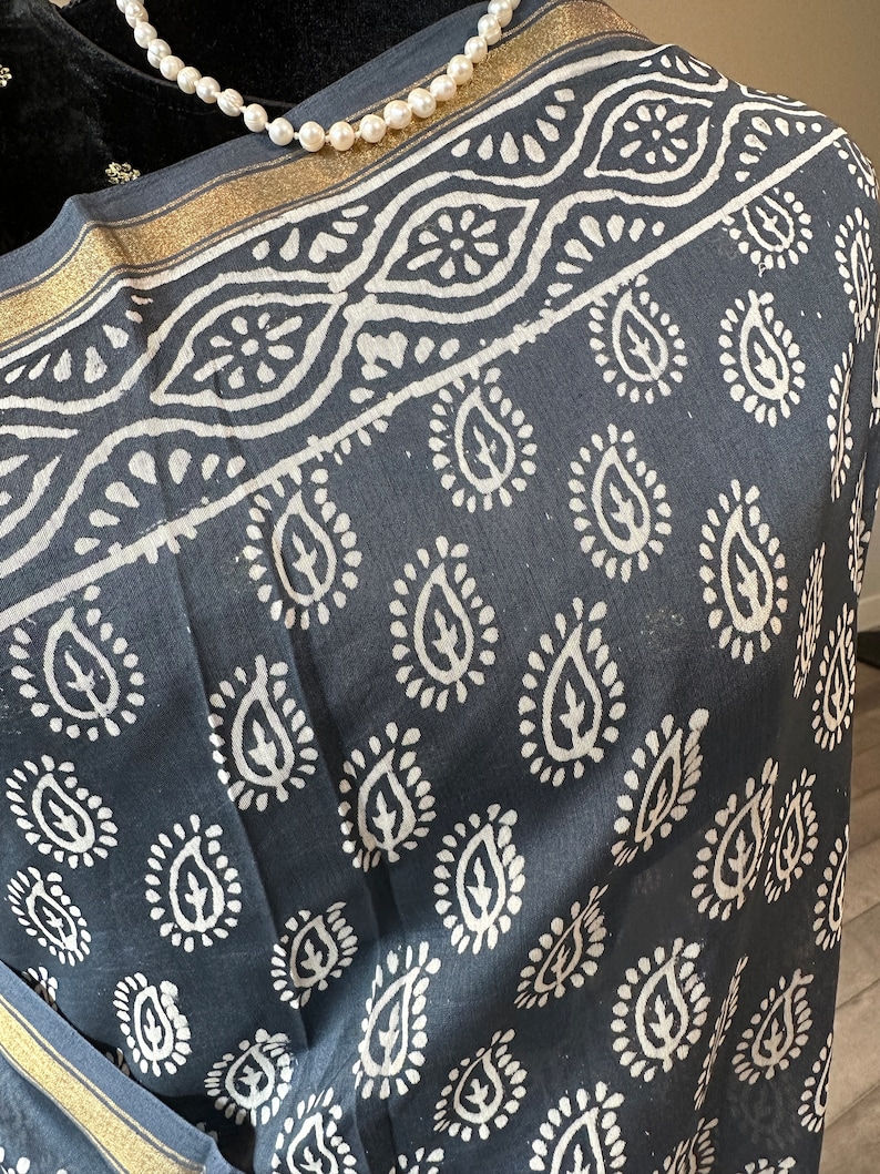 May include: A black cotton scarf with a white paisley pattern and gold trim. The scarf is folded and draped over a black surface.