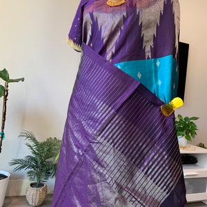 May include: A purple and blue silk saree with silver accents and a gold necklace. The saree has a striped pattern and a border with tassels.