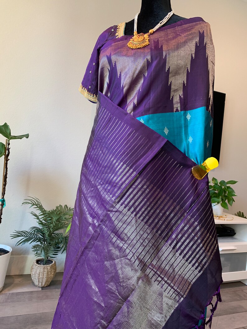 May include: A purple and blue silk saree with silver accents and a gold necklace. The saree has a striped pattern and a border with tassels.