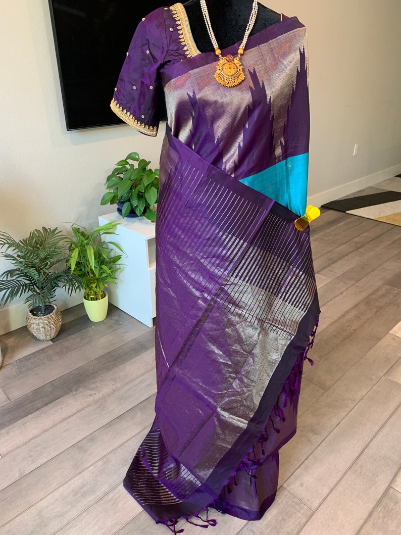 May include: A purple and silver silk saree with a teal blue border and gold trim. The saree has a traditional design and is perfect for special occasions.