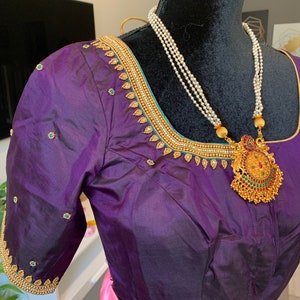 May include: A purple silk blouse with gold and turquoise embroidery. The blouse has a round neckline and short sleeves. A pearl necklace with a gold pendant is worn over the blouse.