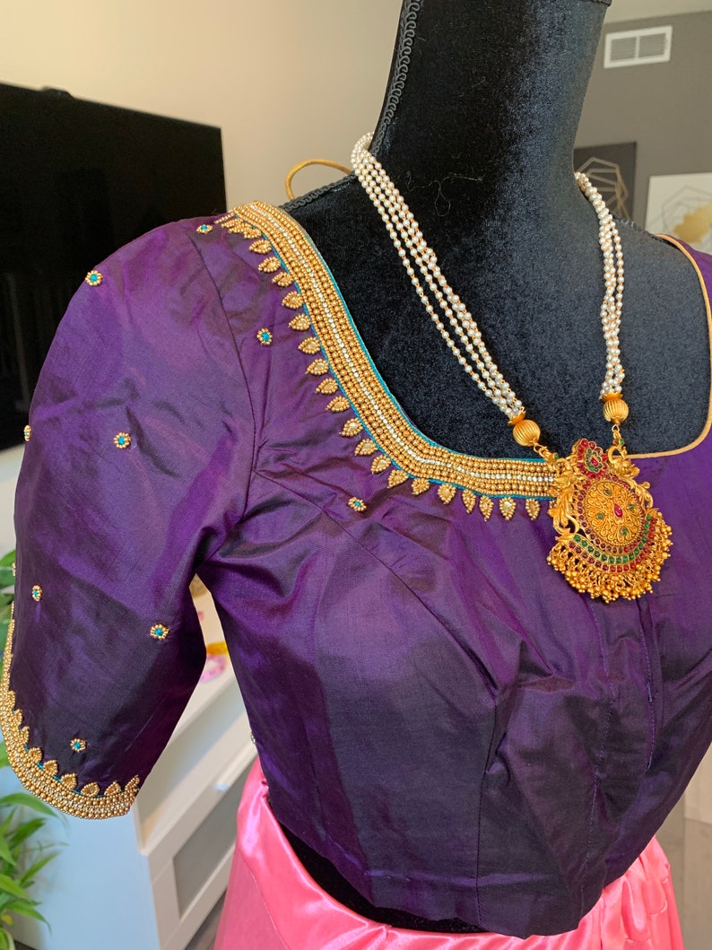May include: A purple silk blouse with gold and turquoise embroidery. The blouse has a round neckline and short sleeves. A pearl necklace with a gold pendant is worn over the blouse.