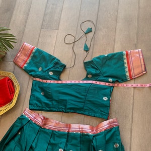 May include: A teal green skirt and matching crop top with a pink and gold trim. The skirt has pleats and the top has short sleeves. The top is measured with a pink tape measure.