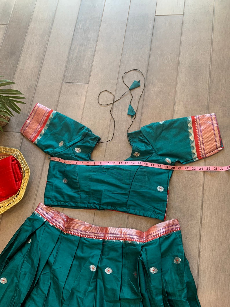 May include: A teal green skirt and matching crop top with a pink and gold trim. The skirt has pleats and the top has short sleeves. The top is measured with a pink tape measure.