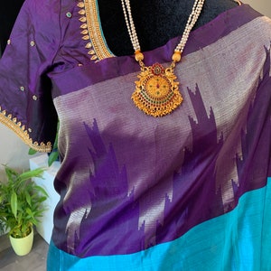 May include: A purple and blue silk saree with a gold and green pendant necklace. The saree has a silver and purple pattern and a blue border.