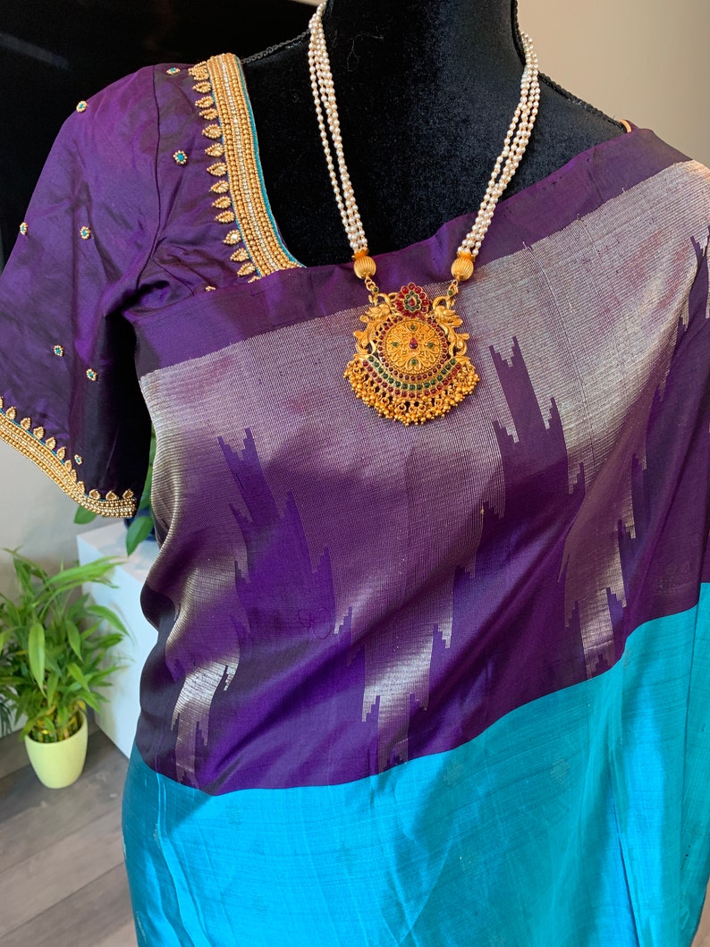 May include: A purple and blue silk saree with a gold and green pendant necklace. The saree has a silver and purple pattern and a blue border.