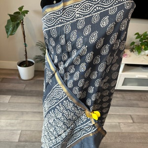 May include: A grey cotton saree with a white paisley print and a gold border. The saree is draped on a mannequin and has yellow clips holding it in place.