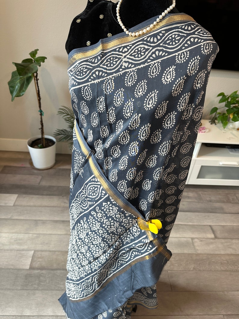 May include: A grey cotton saree with a white paisley print and a gold border. The saree is draped on a mannequin and has yellow clips holding it in place.