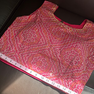 May include: A pink sleeveless top with a gold and white geometric pattern and sequins. The top is measured with a tape measure, showing a length of 17 inches.