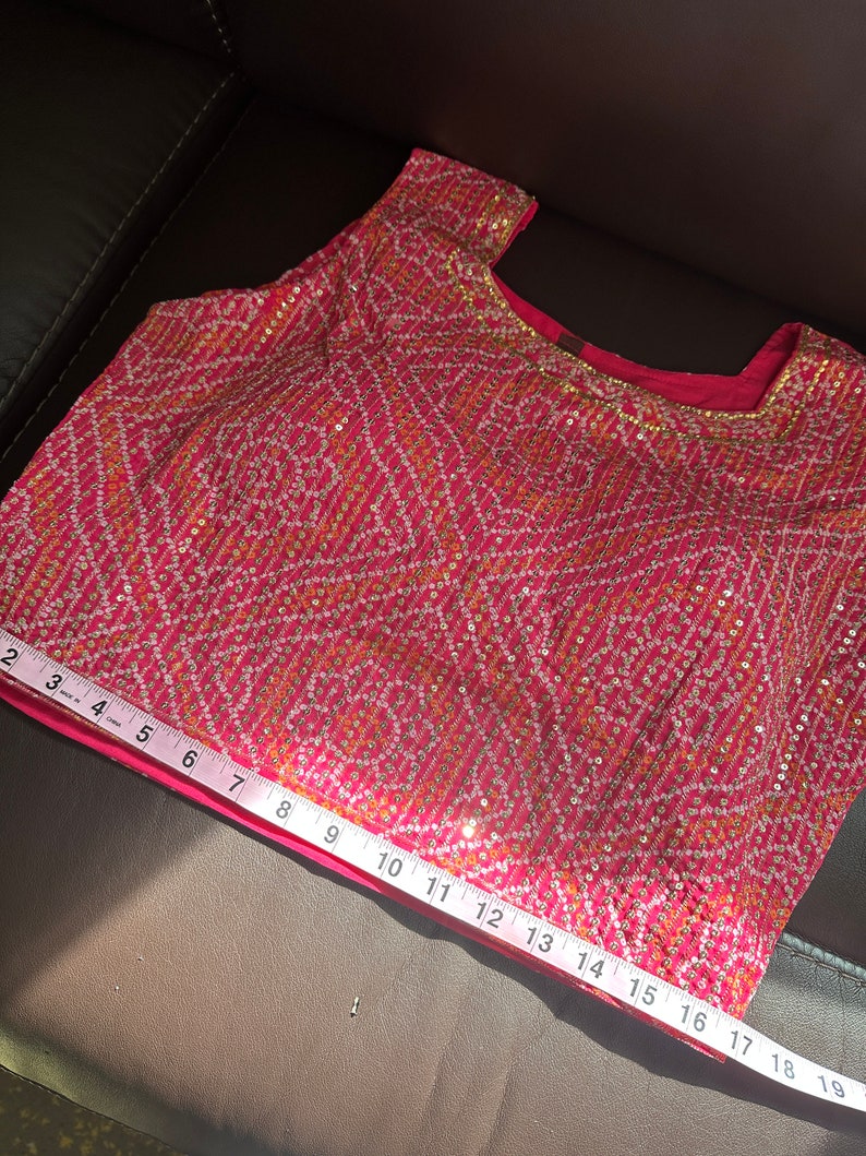 May include: A pink sleeveless top with a gold and white geometric pattern and sequins. The top is measured with a tape measure, showing a length of 17 inches.