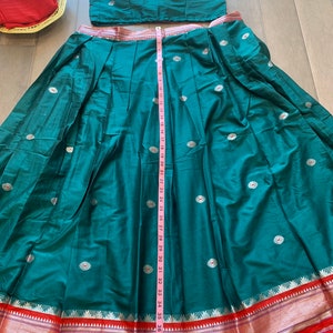 May include: A teal green skirt with a red and gold border. The skirt is pleated and has white floral embroidery. A measuring tape is placed along the side of the skirt, showing a length of 39 inches.