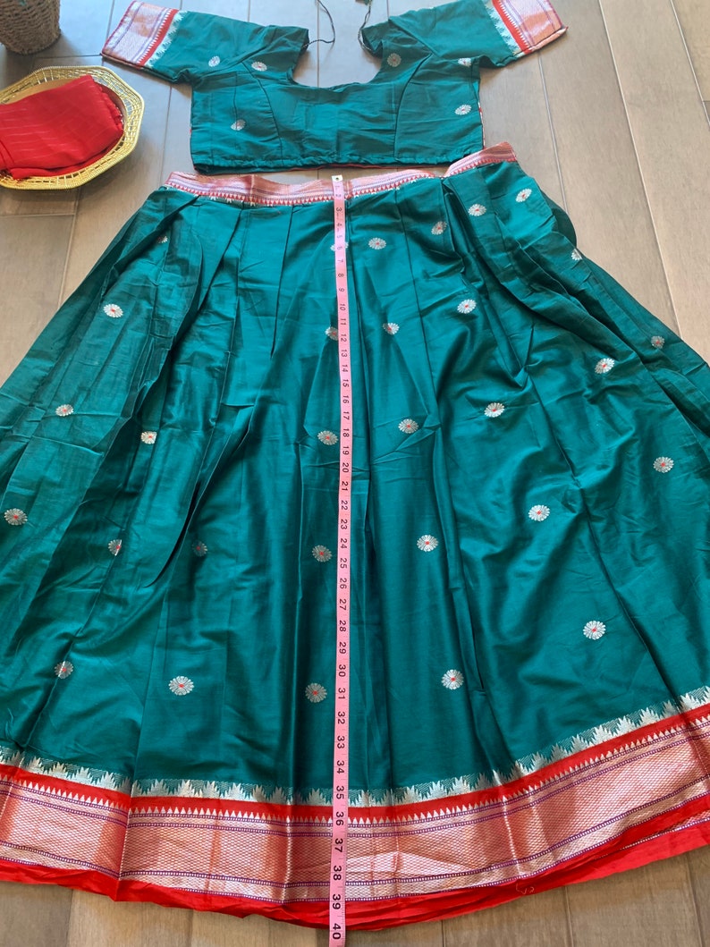 May include: A teal green skirt with a red and gold border. The skirt is pleated and has white floral embroidery. A measuring tape is placed along the side of the skirt, showing a length of 39 inches.