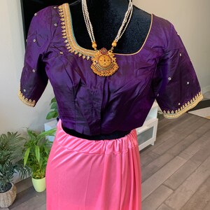 May include: A purple silk blouse with gold and green embroidery and a pink satin skirt. The blouse has a round neckline and short sleeves. The skirt is a simple, flowing style. The model is wearing a gold necklace with a large pendant.