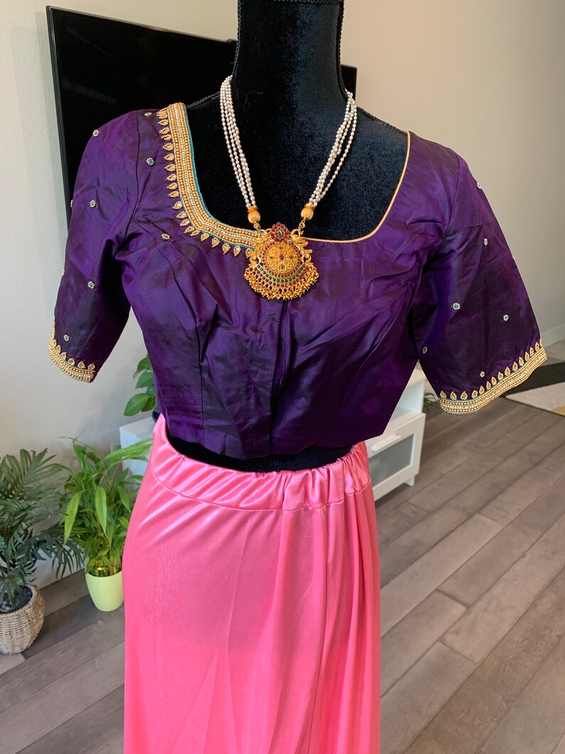 May include: A purple silk blouse with gold and green embroidery and a pink satin skirt. The blouse has a round neckline and short sleeves. The skirt is a simple, flowing style. The model is wearing a gold necklace with a large pendant.