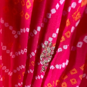 May include: Pink fabric with a white and orange geometric pattern. The fabric has a pleated texture and is decorated with a silver and gold embellishment.