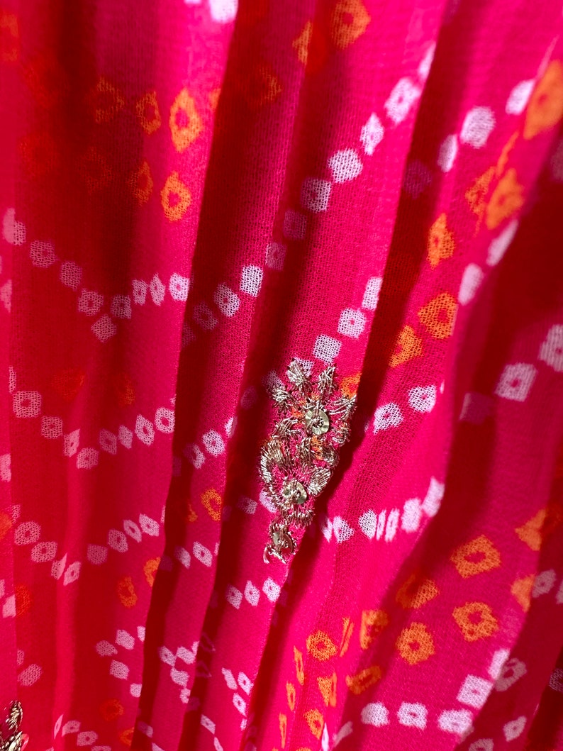 May include: Pink fabric with a white and orange geometric pattern. The fabric has a pleated texture and is decorated with a silver and gold embellishment.