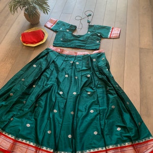 May include: A green skirt with silver floral embroidery and a red and gold border. The skirt is paired with a matching green crop top with short sleeves and a red and gold border.