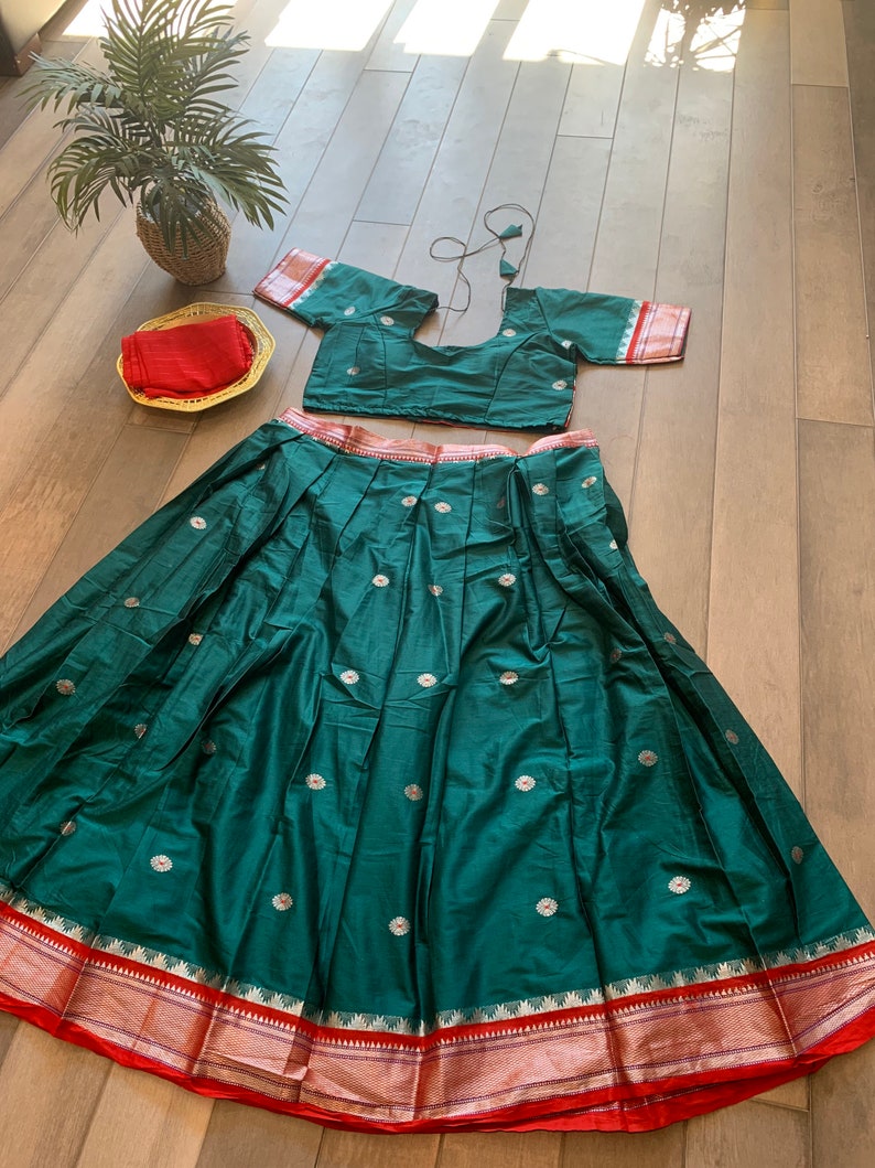 May include: A green skirt with silver floral embroidery and a red and gold border. The skirt is paired with a matching green crop top with short sleeves and a red and gold border.
