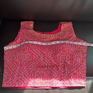May include: A pink sleeveless top with a gold and white embroidered design. The top is measured with a tape measure, showing a length of 18 inches.