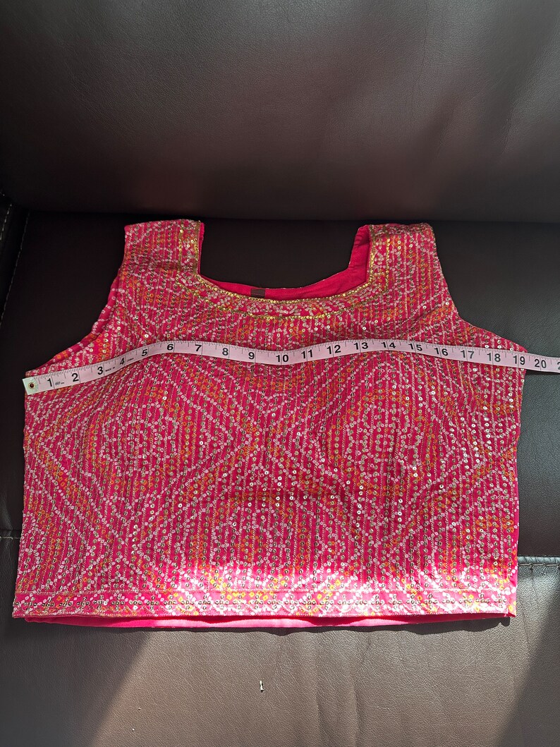 May include: A pink sleeveless top with a gold and white embroidered design. The top is measured with a tape measure, showing a length of 18 inches.