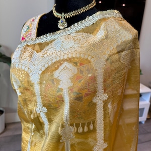 May include: A gold and white saree with a silver border and intricate floral designs. The saree is draped over a mannequin and features a gold necklace with a blue gemstone pendant.